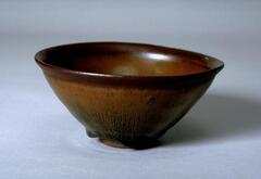 A deep, conical bowl on a straight foot ring with subtle rim articulation.  It is covered in a thickly applied, dark iron-rich black glaze with profuse lighter russet-brown hare's fur (兔毫盏 <em>tuhao zhan</em>) markings.  The thick glaze thins at the rim to a russet-brown color and pools near the foot ring in dark wide drips.