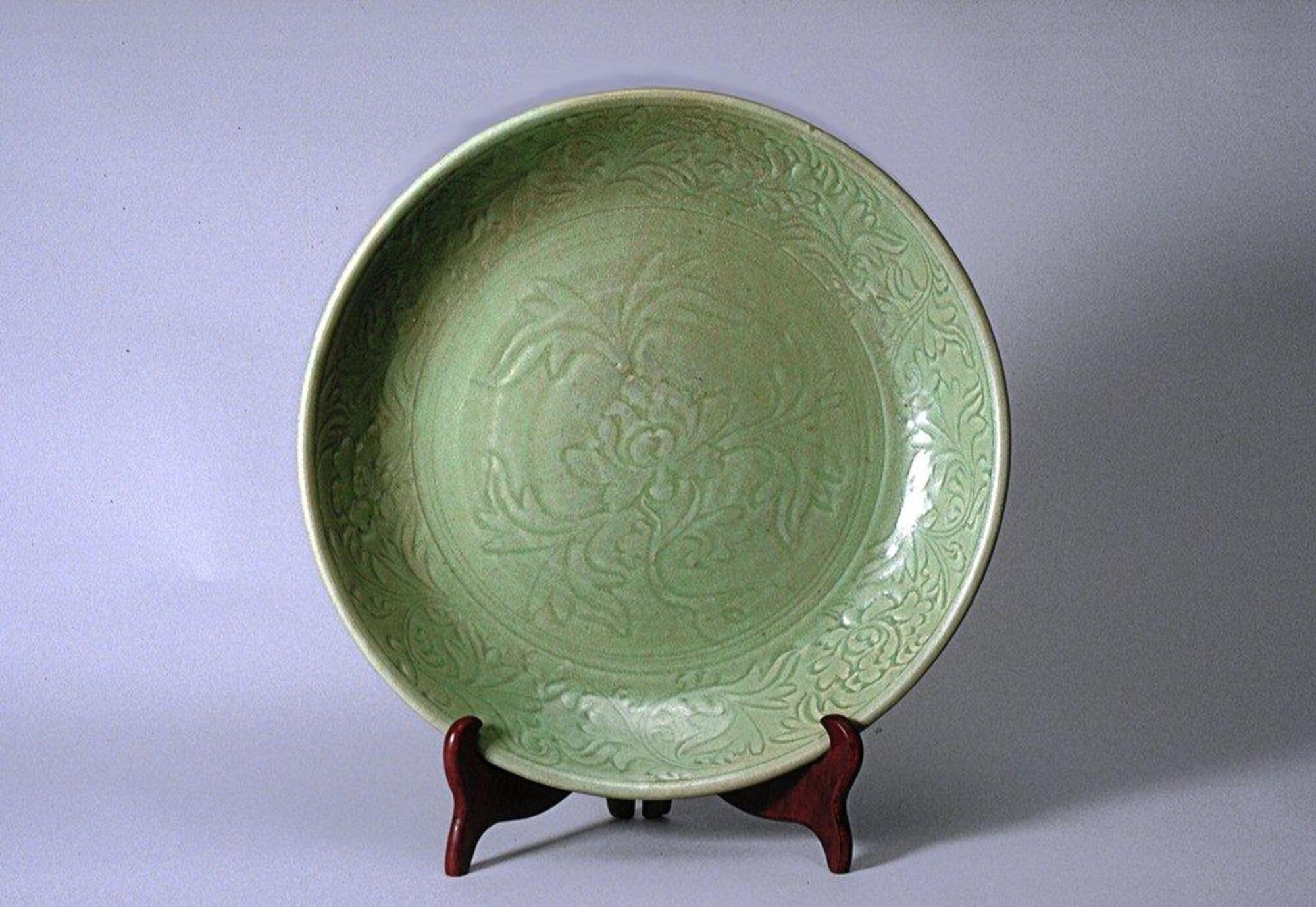 This large stoneware charger has a flat base on a foot ring and short, inverted, gently-curving sides with a direct rim. The interior is incised with a central peony motif surrounded by a floral meander border. It is covered in a green celadon glaze.