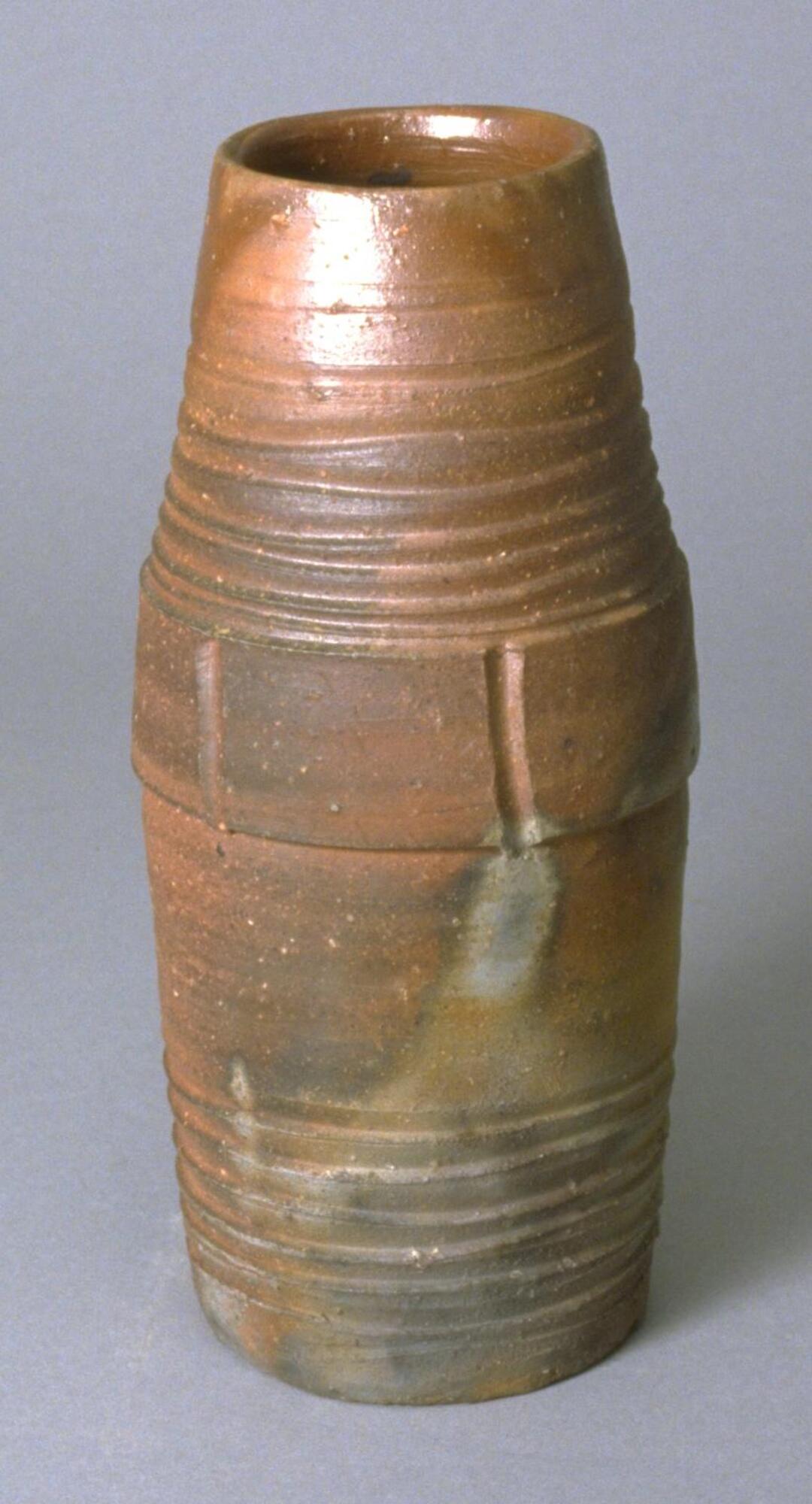 This is a tall, cylindrical vase, slightly bulged at the middle. There are multiple incised lines around the neck and the bottom. The middle part has several vertical incision marks. The bottom is flat. It has a brown body, unglazed, with grayish ash glaze marks.<br />