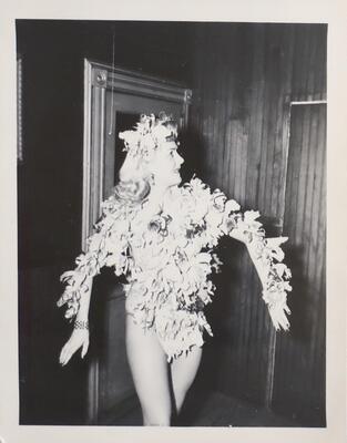 A woman in a wood-paneled room wearing a costume made from what appear to be flowers. The flowers cover her from her neck to her hips and down both her arms, and there are also a few in her hair. She poses with her head to the side and her arms held out from her sides.