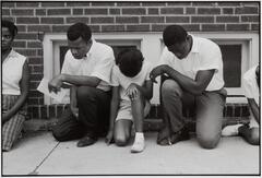 A group of African Americans praying while kneeling on a street. There is concrete pavement and a brick building behind them.