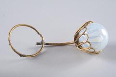 A clouded glass orb in a brass-colored metallic cap comprised of looped petal-like shapes connected to a thin metal stem, which in turn is attached to a thin brass ring about the same size as the glass orb.