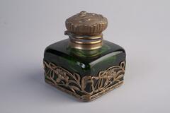 Inkwell made of green glass has a cube shape body and a round metal lid. The body of the inkwell is decorated with brass flower craftings.