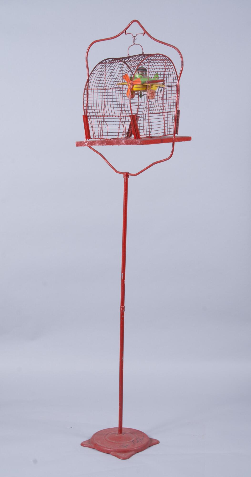This sculpture consists of a red bird cage stand with a half-circle red cage on top. Inside the cage, there is a plastic green, yellow and red airplane toy hanging from the top. There is a white and black figure inside the plane. The work is signed by the artist in black paint at the base: "Tyree Guyton".