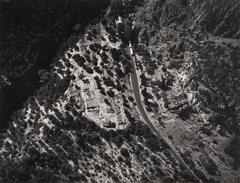 This photograph depicts an aerial view of an archaeological site on a mountain. A road stretches through the mountainside, which is covered in trees and overlooks a coastal plain.