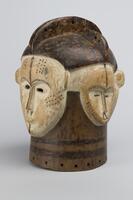 An enclosed wooden mask with four white faces on top of a darker wood. Two faces are larger and two are smaller, all with decorative marks on them. There are holes drilled along the bottom and through the ridges on the top.