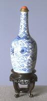 A blue and white snuff bottle in the shape of a vase with a long neck. It is stylized with floral patterns and has a brass collar and coral stopper.