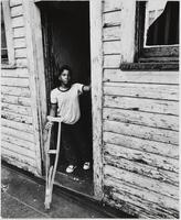 Image of a boy standing in the doorway of a house with peeling, painted wood siding. He looks directly at the camera holding a pair of crutches.