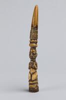 Ivory rod with a round base and a pointed tip. A carved female figure kneels near the bottom of the rod. Above and below her are rows of various carved geometric patterns. A portion of the piece is hollow and there is an intentional hole at the base.