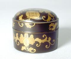 A small cylindrial box with a rounded top. Gold lacquered floral patterns wrap around the entire surface, and a small geometric crest appears in gold on the top of the box. Part of a bridal trousseau.