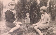 Two children straddling a log with a doll standing on the log between them. The child on the left is looking at the camera and the child on the right is looking down towards the doll.