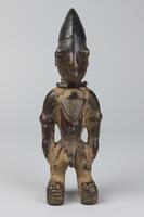 Standing male figure with each foot on a raised base. The hands are at the side and on the front and back of the chest is a triangular pendant with a pattern of raised dashes. There is a string of beads around the neck and on each cheek are three horizontal grooves. The hair is a conical shape. 