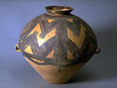 A light reddish-buff earthenware <em>guan (</em>罐) jar with a wide globular upper body and conical lower body on a narrow flat base, and a tall narrow neck with everted rim. There are two diametrically opposed lug handles at the waist. The upper half of the body is painted with black and red pigments to depict two concentric circles on opposing sides. The circles contain network patterns, separating two main motifs of zigzags divided by a central vertical line. Six or seven thin lines extend from each of the points, confined between solid band borders, with a lobed line border below. Around the neck are bold, thick, black, slightly curving brush strokes.