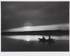 This photograph depicts a silhouette of three figures in a boat paddling across a body of water while the sun sets behind them.