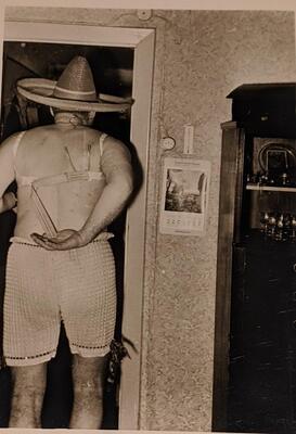 An off-center figure wearing shorts, a brassiere, and a sombrero walks under a doorframe with their back to the viewer. The figure&#39;s right arm is reaching behind their back for a strap on the&nbsp;brassiere.