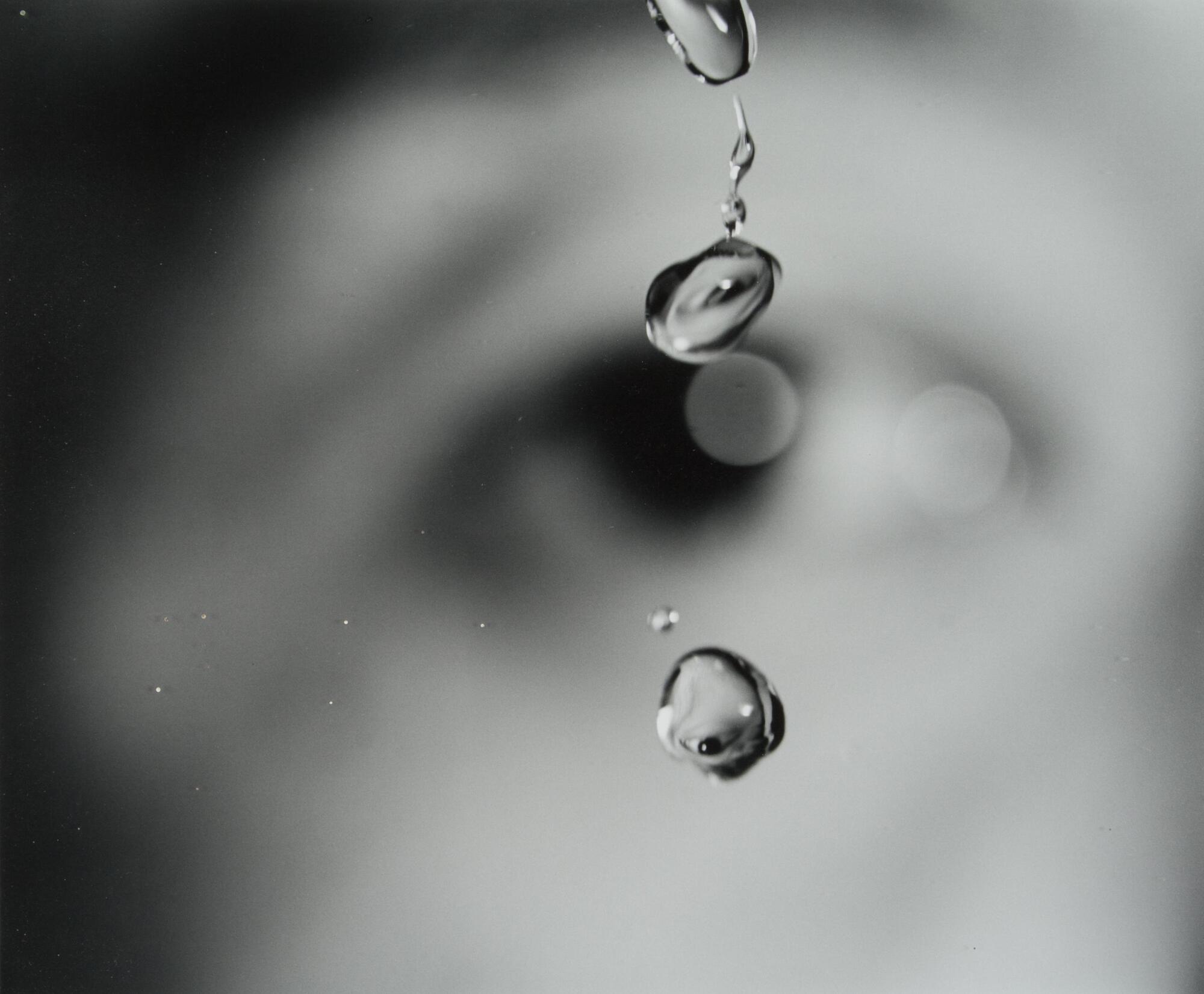 A black and white photograph of droplets of water captured in suspension. Behind the water droplets, an eye is visible, though it is blurred by a shallow depth of field. In the water droplets, the eye is reflected upside down and in sharp focus.