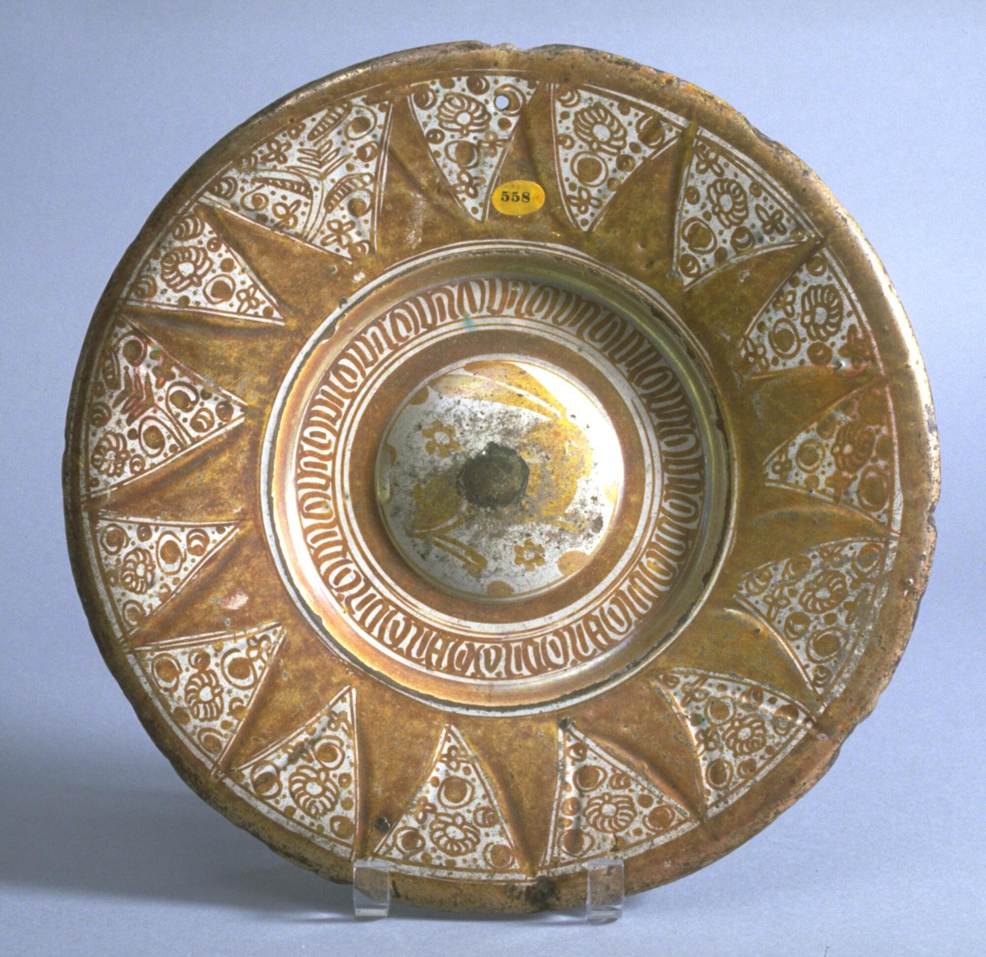 This dish features a three part structural division; the boss is almost flat. Around the depressed area is a band of degenerate Gothic [or pseudo-Arabic] script. On the brim are solid lustre painted zig-zags, possibly a late version of gadroons. The empty areas of this pattern are filled with dots and floral motifs. The reverse has repeated circles only.