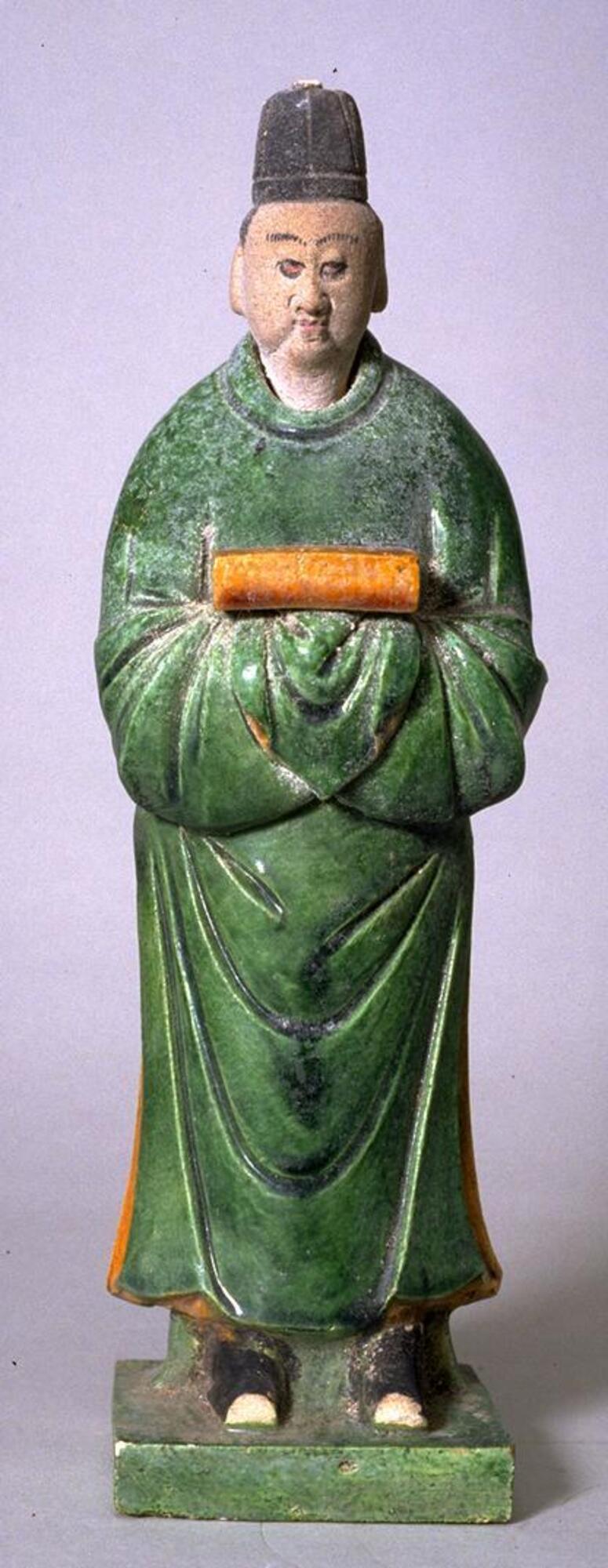 An earthenware male figure standing on a platform wearing long green robes and a tall black hat, holding an amber-colored plaque in his hands, his face painted with polychrome mineral pigments. 