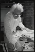 A man with white hair and glasses sorting through a pile of papers on a desk.