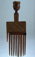 This wooden comb includes 11 large teeth, one of which has broken off. Its handle includes a rectangular section with multiple bands of diagonal, incised lines. This rectangular section along with the teeth of the comb visually form an abstracted body for the delicately carved head, which sits atop the handle. The head itself bears fine facial features as evidenced by its straight mouth and narrow eyes with large, round orbits.  