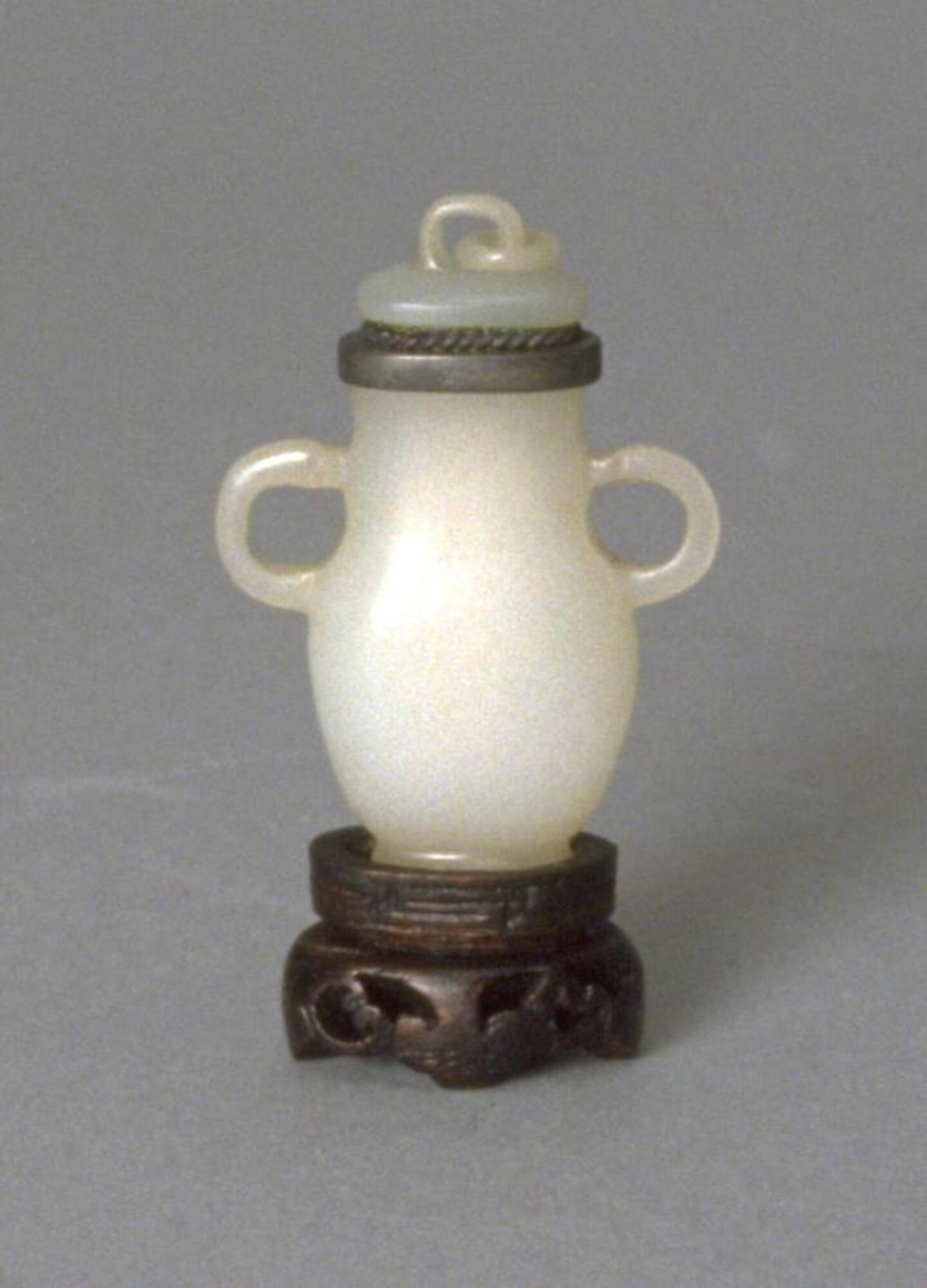 A white nephrite jade snuff bottle, shaped like a vase with two large circular handles on the side. The snuff bottle has a silver colored rim, with a stopper that also has a circular handle.&nbsp;