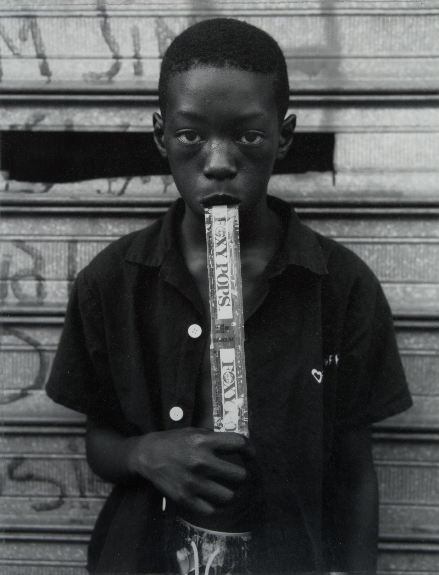 A photograph of a young African American boy standing against a metal slatted wall, on which graffiti is visible. He wears a dark button down shirt and holds a Foxy pop in his hand, eating it as the photograph was taken.