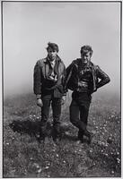 A photograph of two men in jackets and jeans standing in a grassy field filled with dandelions. Behind them, a foggy atmosphere obscures the background. They stand side by side, looking toward the camera.