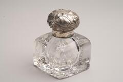This is a cut crystal inkwell with a sterling silver collar and cap. It has a square shape with smooth rounded edges at the top and a geometric cut design on the bottom. The round shaped lid has vegetal designs.<br />