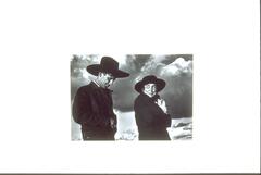 This photograph depicts a man and a woman wearing black cowboy hats and dark jackets and framed against a cloudy sky.