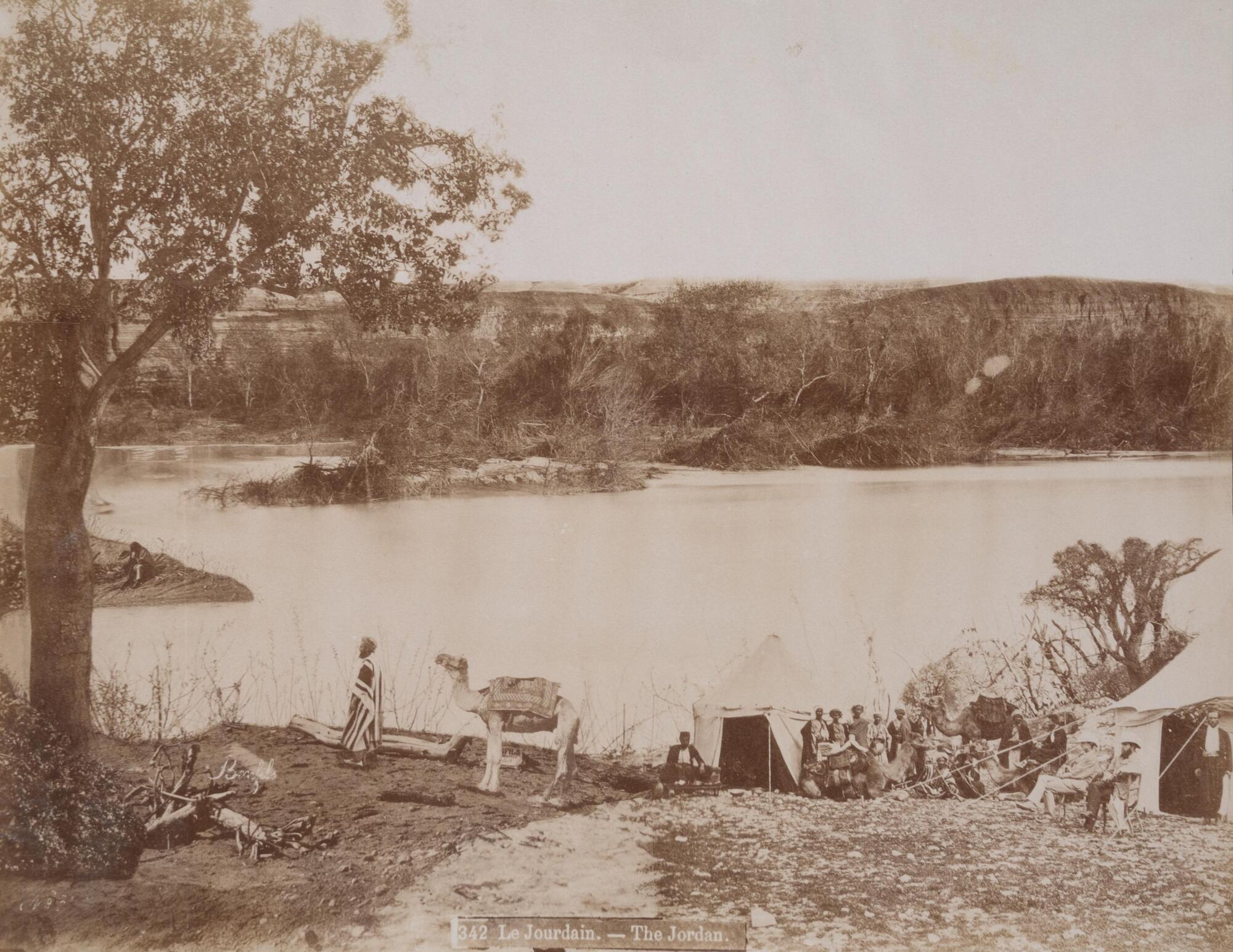 In this photograph, a group of men sit or stand near two tents along the bank of a wide river. The hills and trees on the opposite bank form the horizon line.