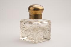 This is a cut crystal inkwell with a brass collar and cap. The bottle is square shaped with starburst designs incised on the corners. The cap has a rounded shape.