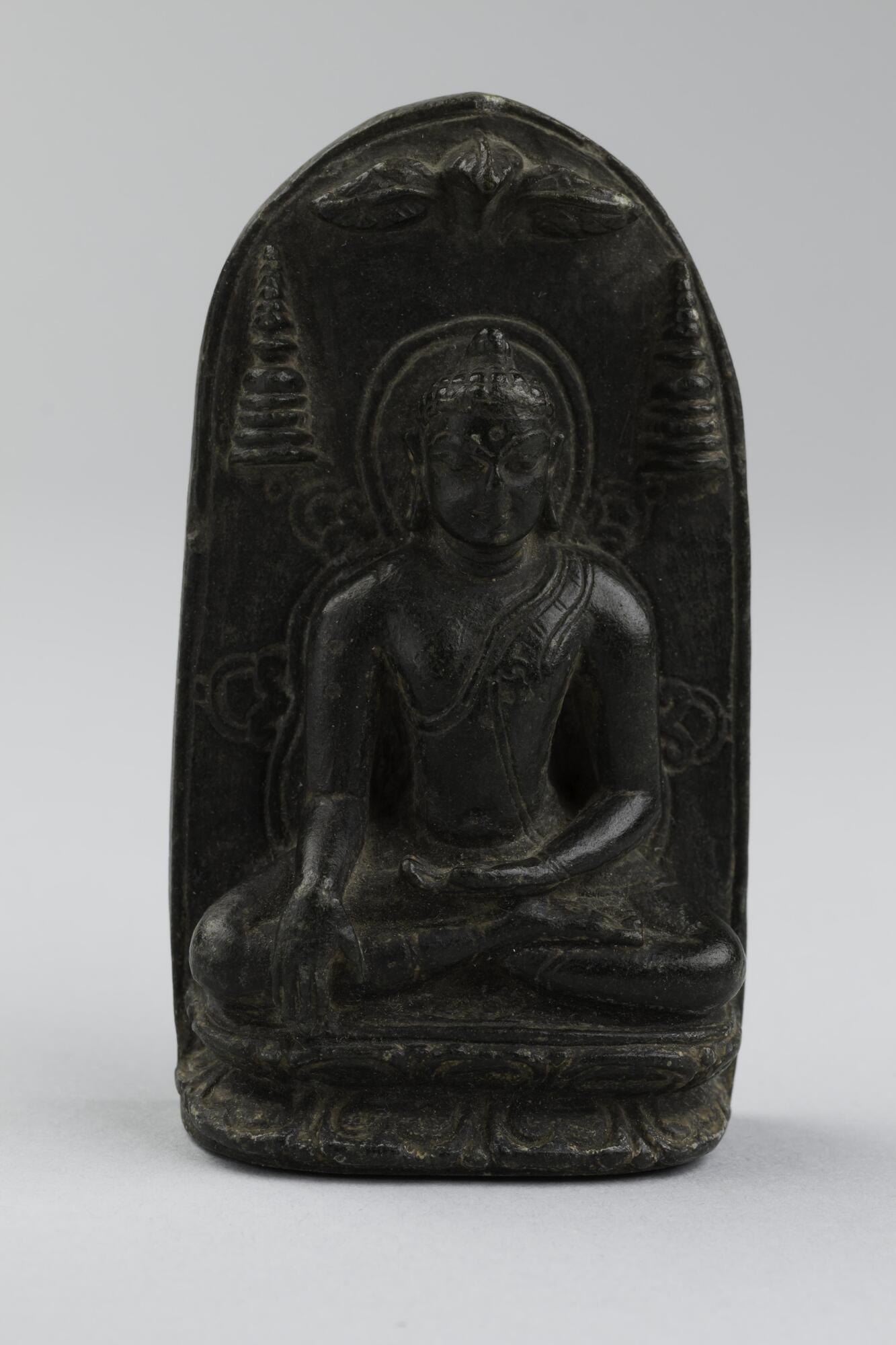 The Buddha in bhumisparsa mudra (the gesture of touching the earth with his right hand, palm inward), signaling his victory over Mara. He is shown under three leaves, indicating the bodhi tree under which he sat while meditating before reaching an awakening. He is shown flanked by two stupas, or reliquary monuments, symbolizing his attainment of nirvana.<br />