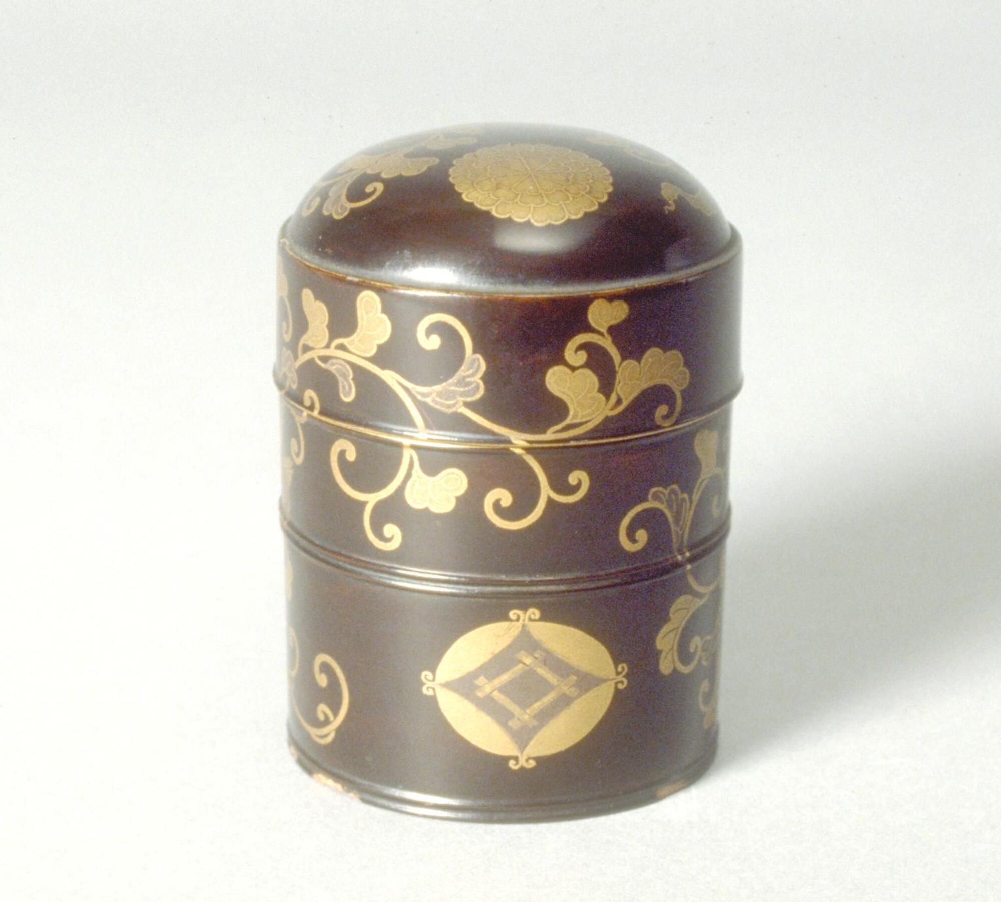 Small cylindrical container lacquered dark brown with gold floral patterns wraping around the object on all sides. There is also a chrysanthemum crest in gold on the rounded top of the container and a geometric crest near the bottom. This is the smaller of two containers. Part of a bridal trousseau.