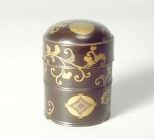Small cylindrical container lacquered dark brown with gold floral patterns wraping around the object on all sides. There is also a chrysanthemum crest in gold on the rounded top of the container and a geometric crest near the bottom. This is the smaller of two containers. Part of a bridal trousseau.