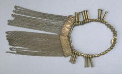 Apron in the form of a metal chain fringe attached to two rectangular brass plates. The apron is attached to a belt of brass beads and pendants that resemble the heads of nails. 