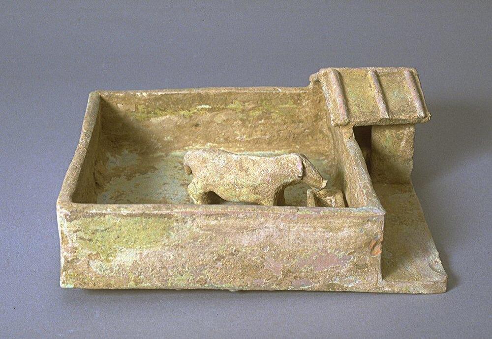 This red earthenware model of a pigpen is square, contains one pig at a feeding trough, and has a peaked roof shed to the side. The model is covered in a green lead glaze.