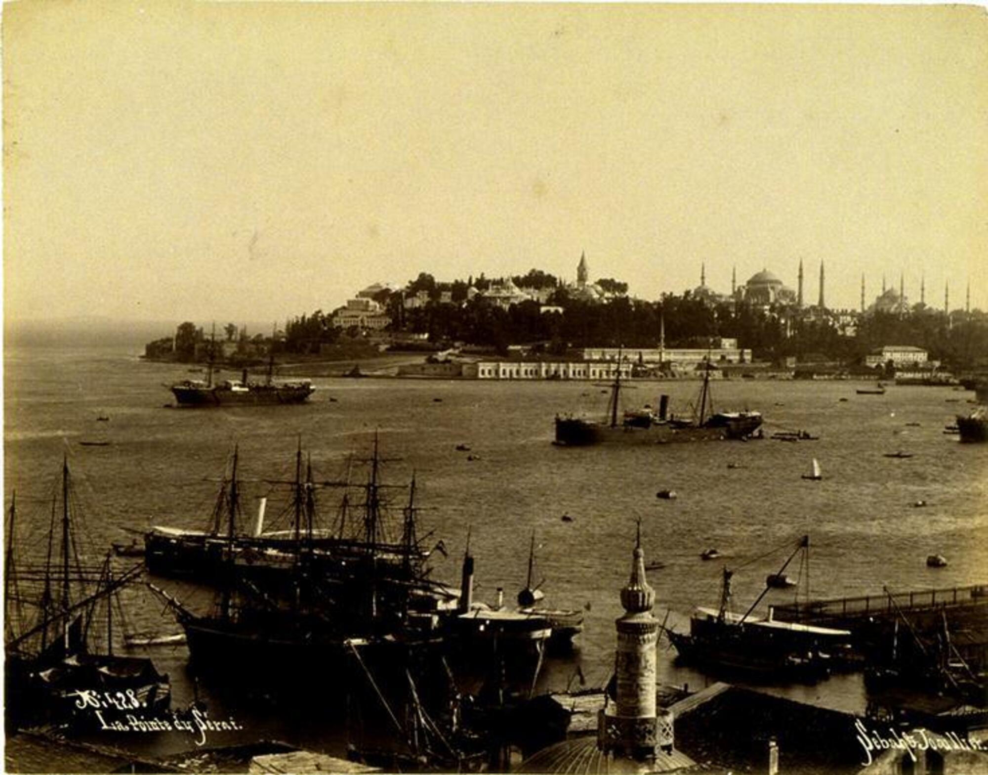 View down onto a busy seaport populated by numerous steamships and smaller watercraft. A sliver of land with various towers and domed structures stretches across the horizon line in the distance. 