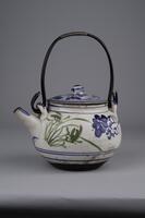White ceramic teapot with a thin black handle and lid. There are patterns of blue flowers on the pot and lid. Patterns of green stems on one side of the pot.