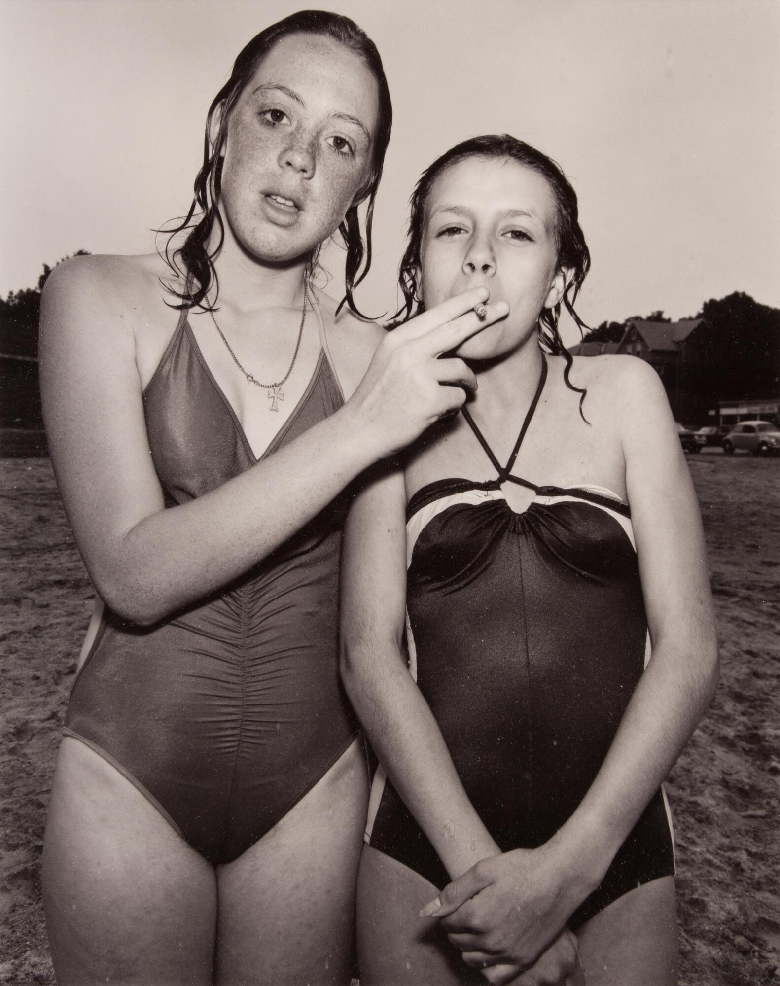A photograph of two girls standing in their bathing suits, their hair wet. The girl on the left wears a celtic cross around her neck and holds a cigarette to the other girl's mouth. 