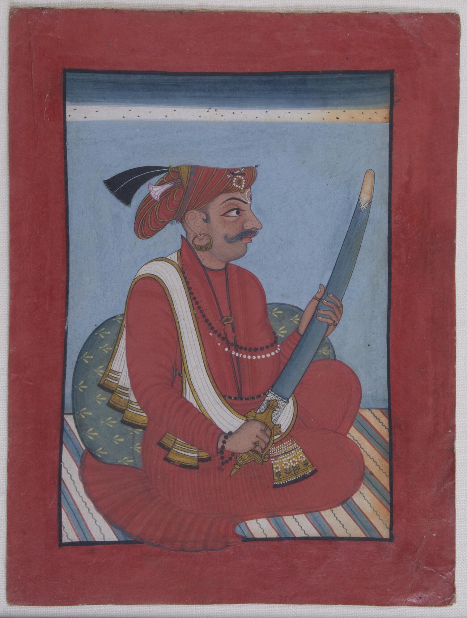 A man sits on a striped surface with his legs crossed under his red clothing. He wears a white sash across his right shoulder and a red hat. In his hands he holds a sheathed sword.