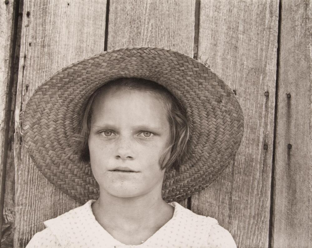 The photograph is a portrait of a girl in a straw hat standing in front of a wooden fence.