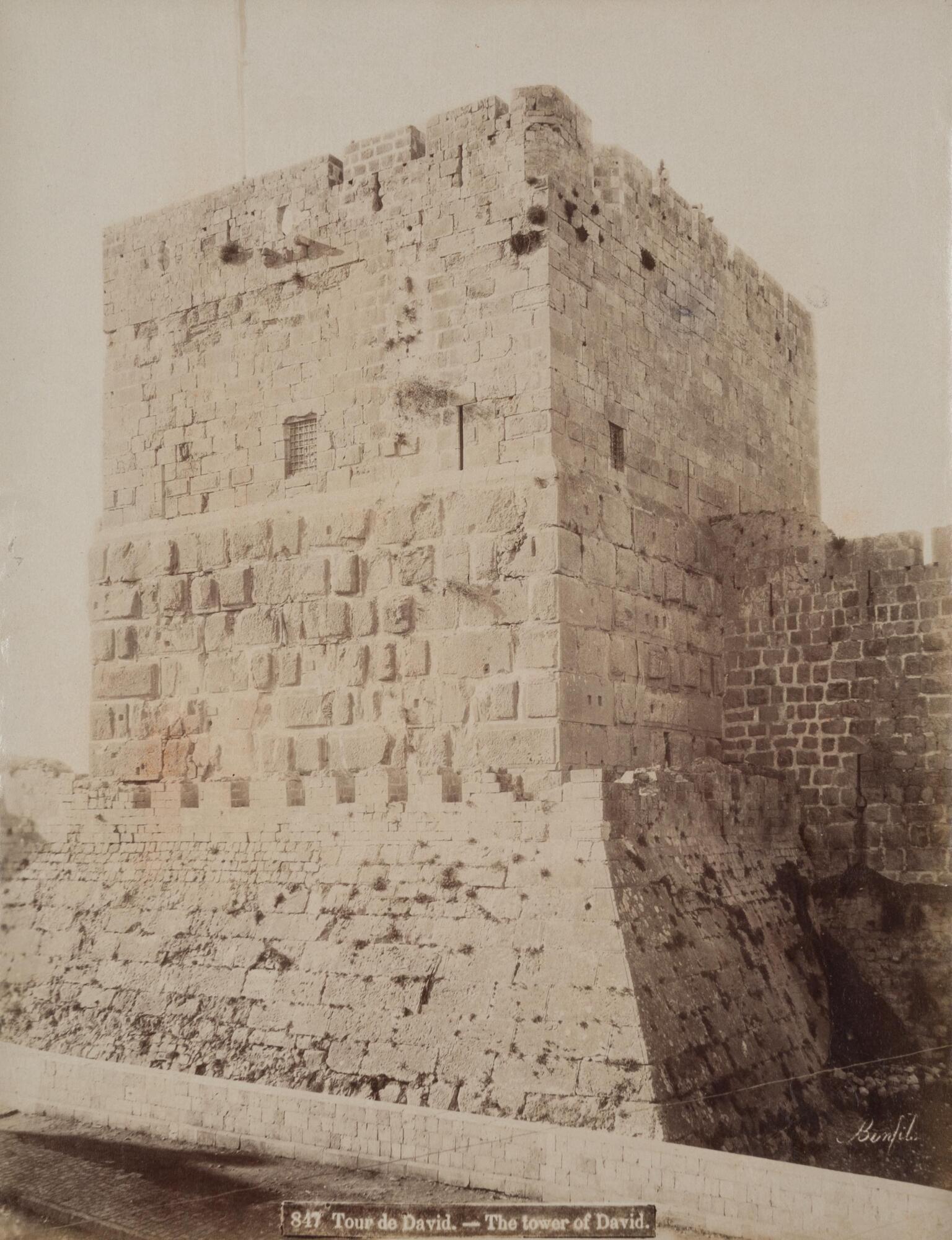 The photograph shows a rectangular stone tower from below with crenellated battlements, small grated windows, and a sloping base.