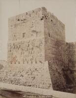 The photograph shows a rectangular stone tower from below with crenellated battlements, small grated windows, and a sloping base.