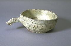 A red, ceramic, shallow, flat-bottomed earthenware bowl.  It has a short dragon handle to one side, and is covered in a green lead glaze with iridescence and calcification. 