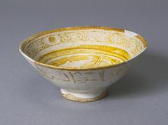 The interior of this bown has two zones of decoration: one just below the rim and the other occupying the remainder of the vessel's surface. The rim area has a series of six solid lustre circles, each surrounded by a white zone and separated from the others by a band composed of dots connected by curving lines which suggest a loose scroll decoration. The remainder of the bowl is filled with the stylized figure of a bird shown in profile and executed in reserve against a solid lustre background. The curve of the bird's body echoes the shape of the bowl creating a visual rhythm which moves clockwise from its head through its breast and upturned wing. The tip of the wing touches the end of the bird's long beak and the circular movement is continued by a leaf which hangs from the beak. Originally, the bird's feathers were indicated by a series of fine lines and embellished with dots but now only vestiges of this decoration remain. 
