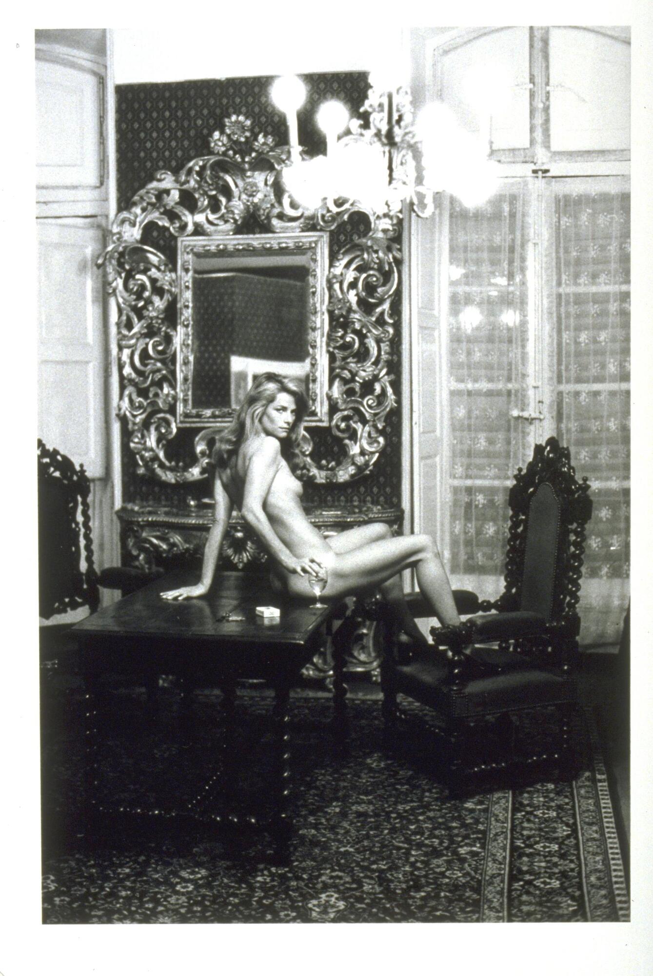 A photograph of a woman seated on a large wood table. Nude aside from a pair of heels, she turns to look at the viewer while resting on arm on the table and the other on a wine glass. 