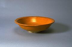 This is a wide, shallow, rounded bowl with everted flat rim on a foot ring, covered in an amber glaze.