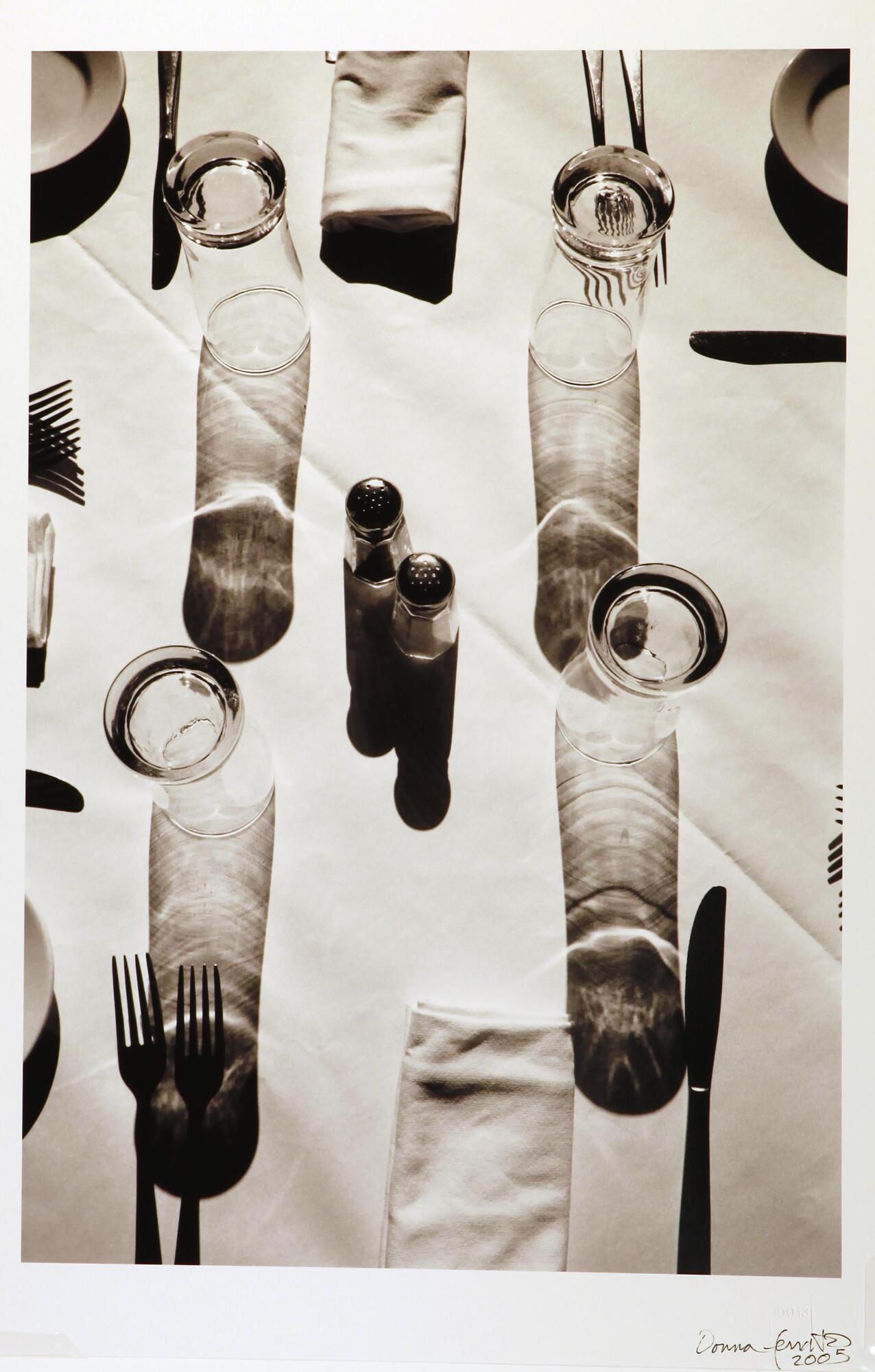 This is a black and white photograph of several place settings on a cloth covered table. Plates, forks and napkins are arranged around salt and pepper shakers in the center. There are four clear glasses that have shadows projecting designs onto the tablecloth. The viewpoint is looking down from above.