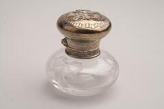 This is a clear cut crystal inkwell with a metal collar and lid. The glass body has a round shape. The lid is also round with a ribbon design and small turquoise stone on top.<br />