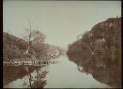 This photograph depicts a view of a lake with a rocky and hilly shoreline, with a small pier that leads into the water. The lake’s surface is quite still and reflects the trees and hills abutting it.  
